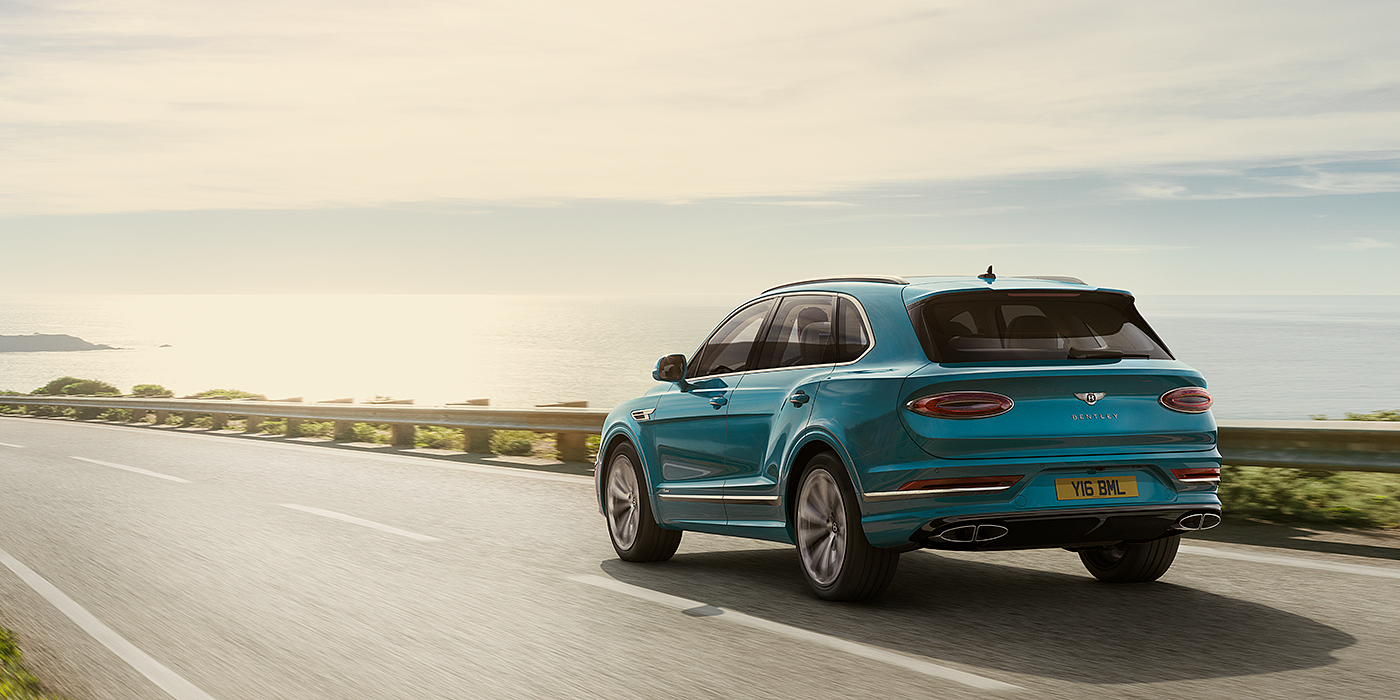 Bentley Doha Bentley Bentayga Azure luxury SUV rear three quarter in Topaz Blue by Mulliner paint, driving on an ocean road.