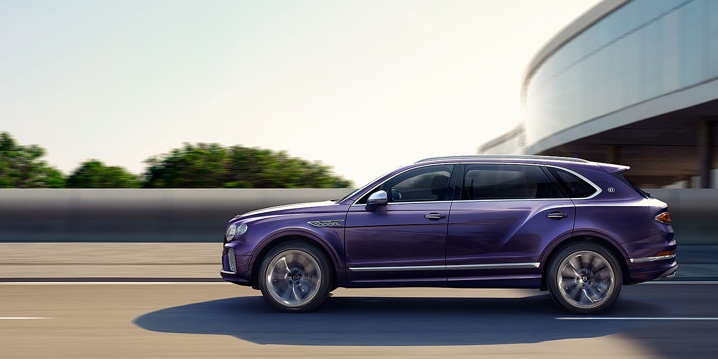 Bentley Doha Bentley Bentayga Extended Wheelbase Mulliner luxury SUV profile in Tanzanite Purple paint, driving on an country road past trees.