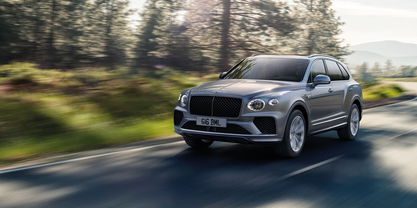 Bentley Doha Bentley Bentayga luxury SUV front three quarter in Moonbeam paint, driving on an country road past trees.