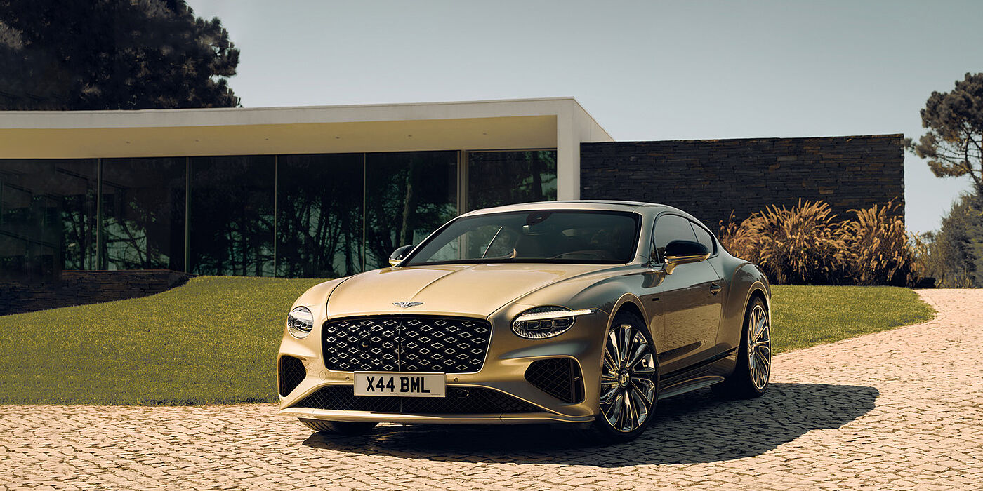 Bentley Doha Bentley Continental GT Mulliner coupe front three quarters in Patina paint parked outside a glass fronted building with a grass lawn behind it