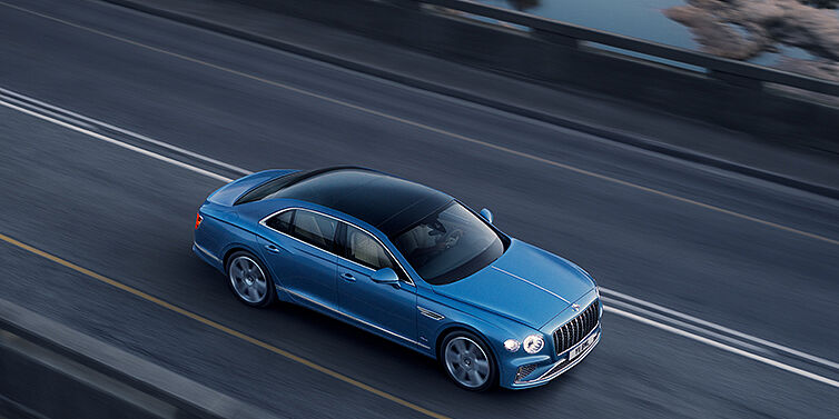Bentley Doha New Bentley Flying Spur Azure sedan in Light windsor blue paint by Mulliner, with 22 inch Azure wheels seen from overhead driving dynamically on a country road