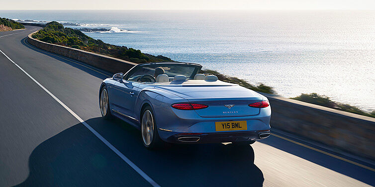 Bentley Doha New Bentley Continental GTC Azure convertible rear view in Light Windsor blue paint with 22 inch ten spoke wheels driving dynamically on an ocean road in the sunshine